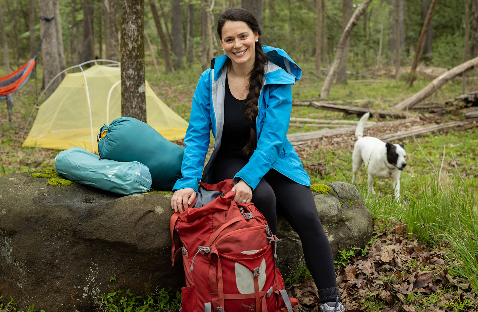 Rada Petric sits on a rock at a campsite with her hands resting on a hiking backpack. A beautiful white dog is in the background.