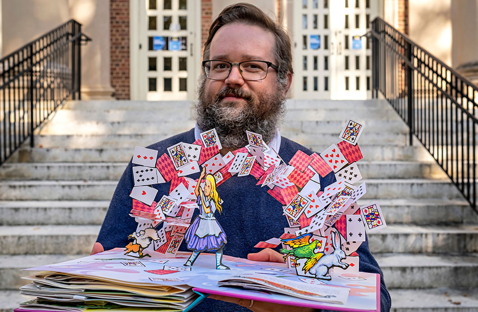 Kenny Jones holding an open pop up book of Alice In Wonderland showing an arch of flying cards and a defensive Alice
