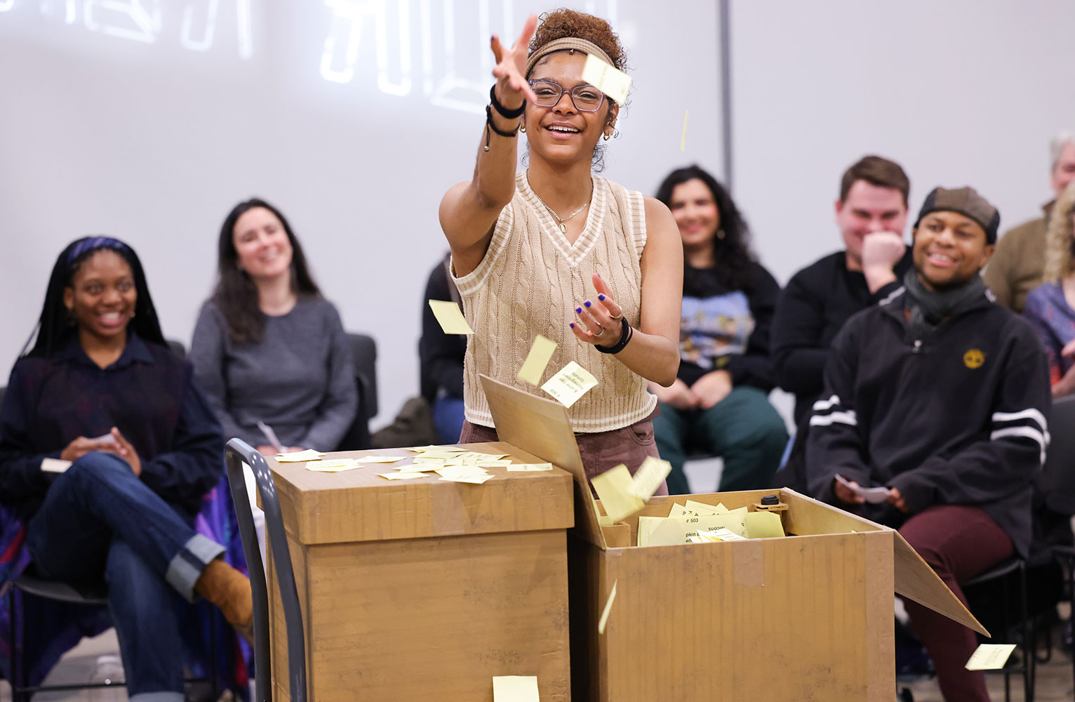 Saleemah Sharpe stands behind two boxes while throwing sticky notes in the air during a performance of Every Brilliant Thing.